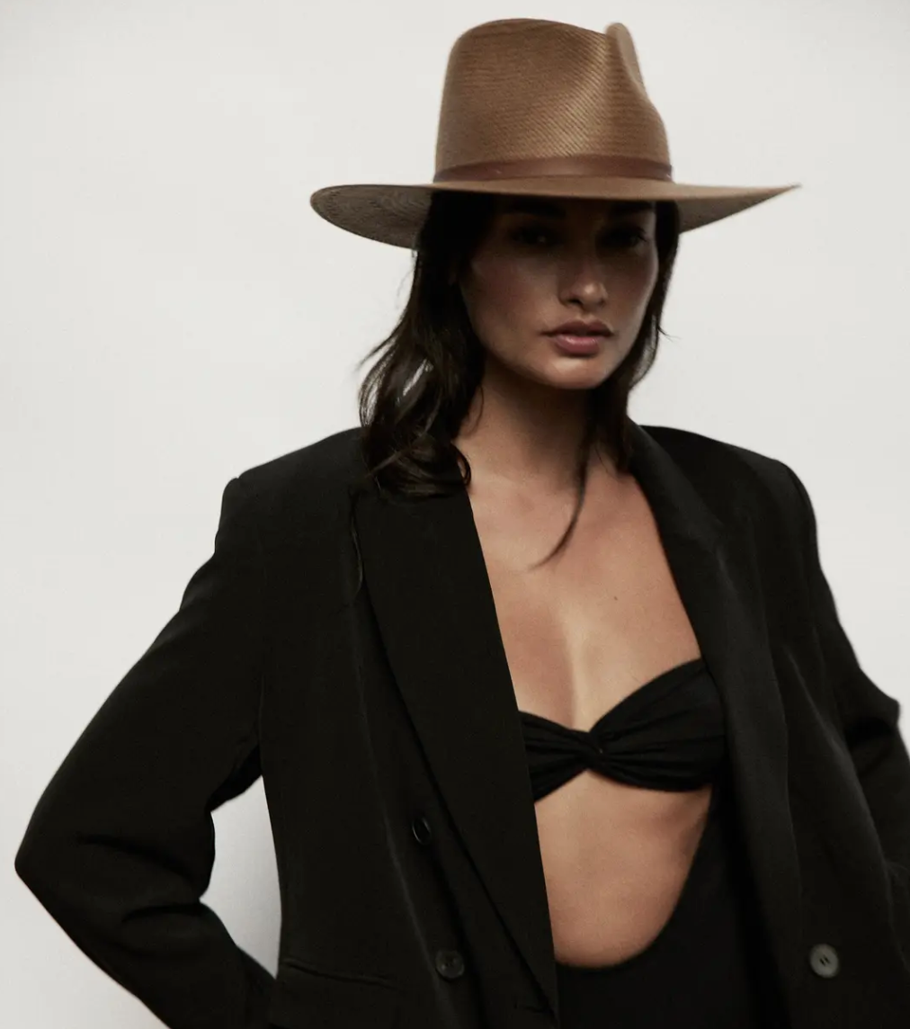 A stylish woman wearing a dark blazer and black bikini top, accessorized with a wide-brimmed Sherman Hat Brown by Janessa Leone, poses against a plain light background. Her Arizona-style expression is confident and subtly intense.