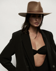 A stylish woman wearing a dark blazer and black bikini top, accessorized with a wide-brimmed Sherman Hat Brown by Janessa Leone, poses against a plain light background. Her Arizona-style expression is confident and subtly intense.