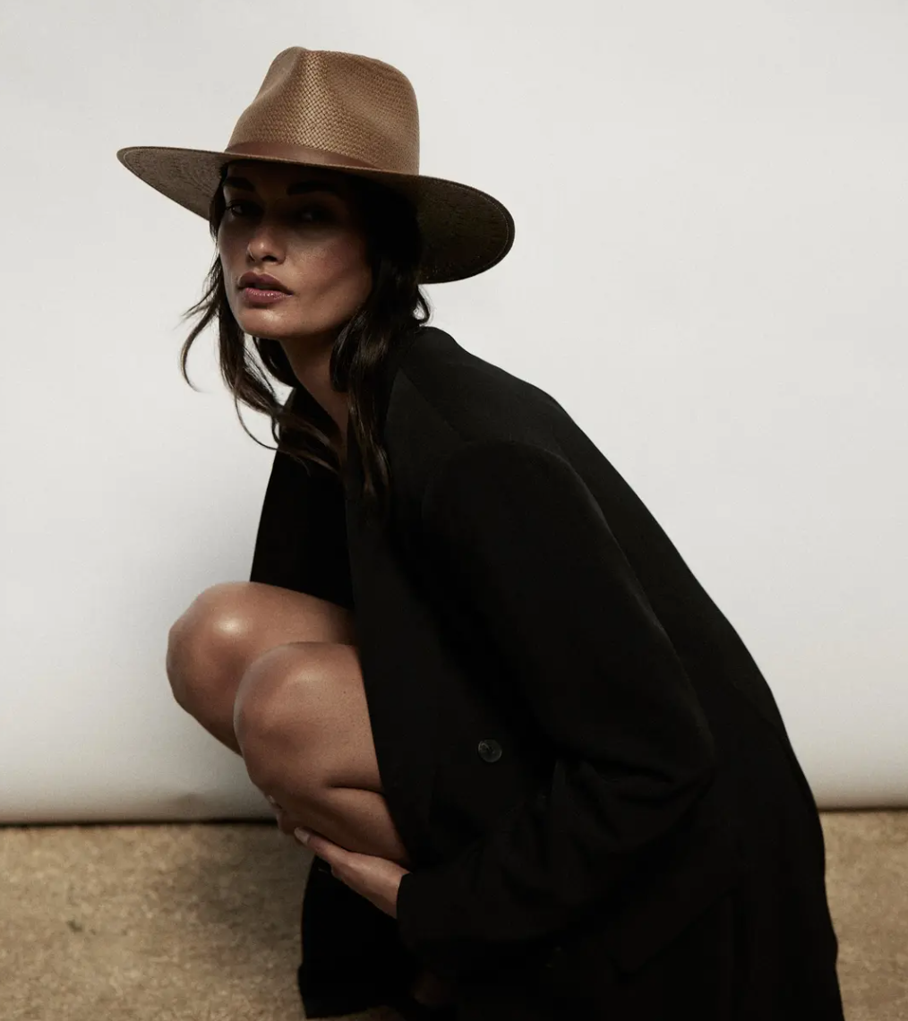 A woman in a stylish black blazer and a tan Janessa Leone Sherman hat kneels, looking intensely at the camera against a plain light background.