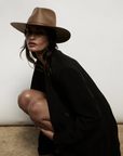 A woman in a stylish black blazer and a tan Janessa Leone Sherman hat kneels, looking intensely at the camera against a plain light background.