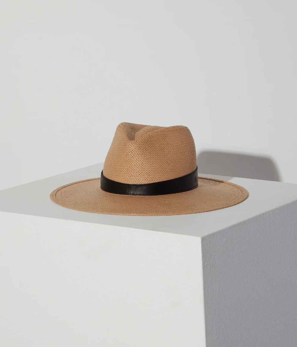 A Savannah hat sand by Janessa Leone with a black ribbon rests on a gray surface against a white background, showcasing its timeless style, with a soft shadow cast on the wall.