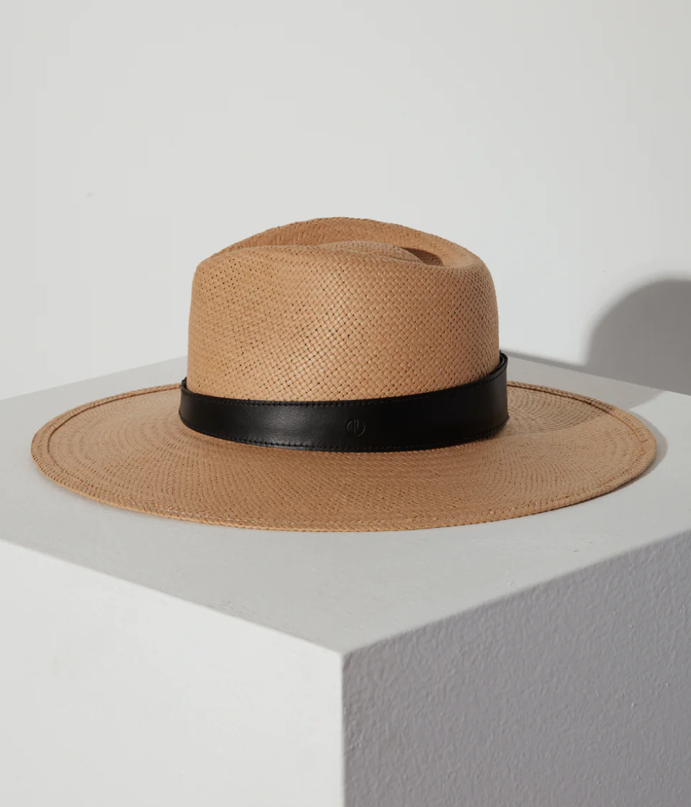 A stylish SAVANNAH HAT SAND by Janessa Leone, with a black ribbon around the base, displayed on a white pedestal against a neutral background with a shadow cast on the wall in an Arizona bungalow.