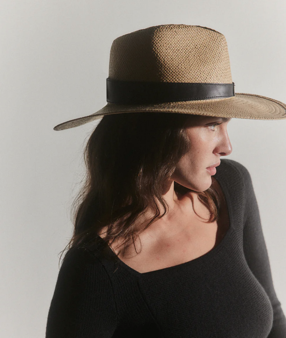 A woman in profile wears a stylish Savannah Hat Sand by Janessa Leone with a black ribbon and a black top, against a light gray background influenced by Arizona style.