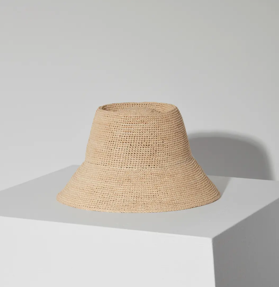 A Felix Bucket Natural hat by Janessa Leone rests on a white cube under soft lighting, casting a gentle shadow to the right, against a plain white background.