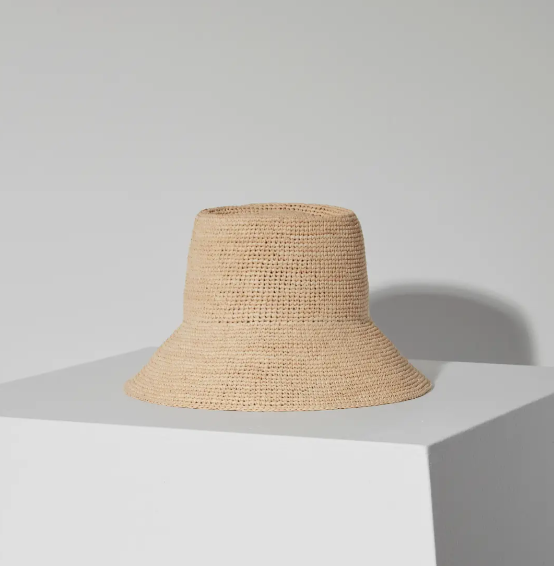 A beige straw Felix Bucket Natural hat in Janessa Leone style displayed on a white pedestal against a plain light grey background, illuminated by soft lighting.
