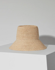 The FELIX BUCKET by Janessa Leone, a beige raffia straw bucket hat, is displayed on a white geometric pedestal against a plain light gray background.