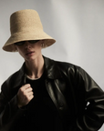 A person wearing the Janessa Leone FELIX BUCKET raffia straw hat, black sunglasses, a black shirt, and a black leather jacket poses against a light, neutral background.