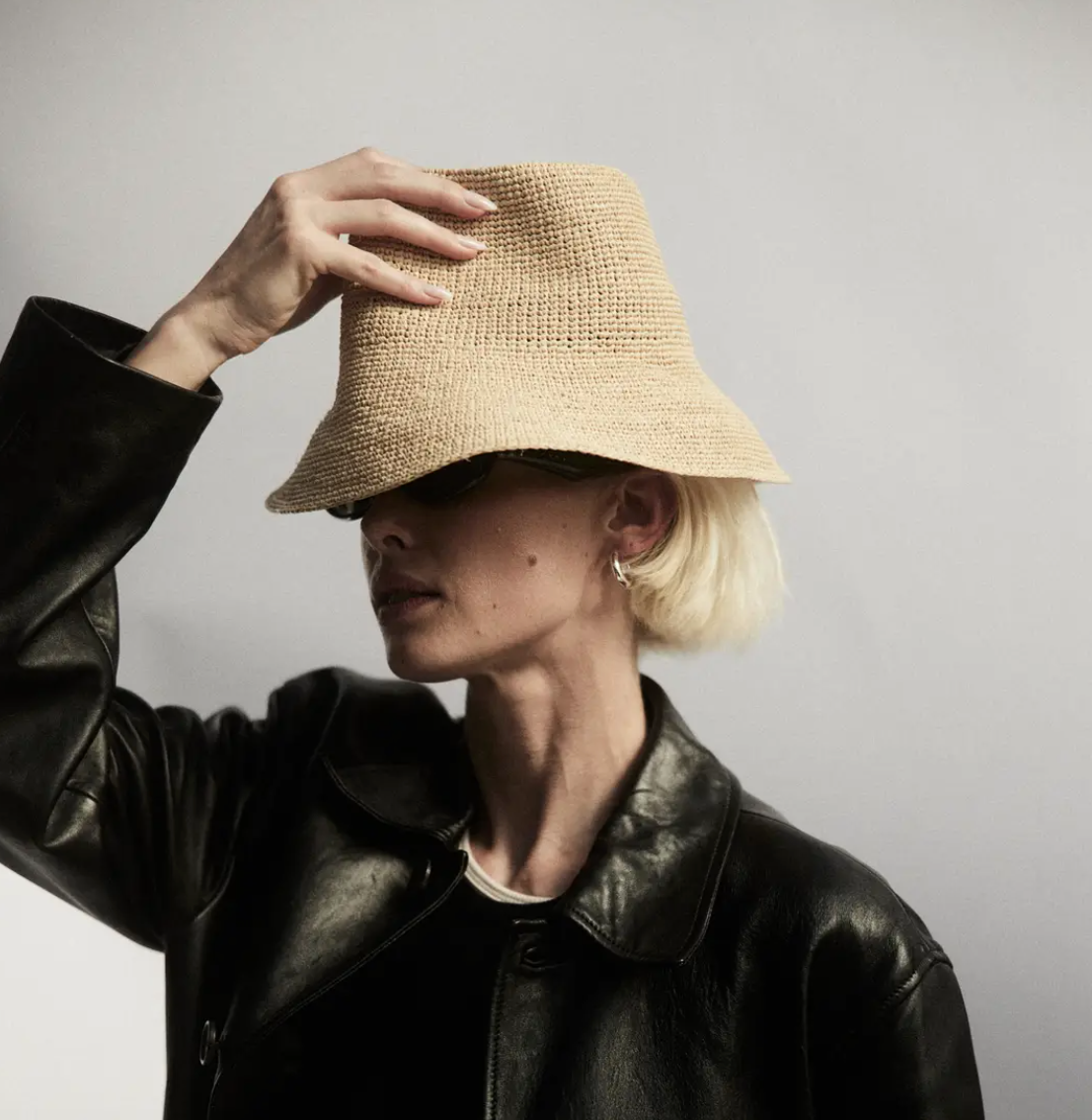 A person in a black leather jacket wearing a wide-brimmed straw hat (FELIX BUCKET NATURAL by Janessa Leone) that covers their eyes, set against an Arizona-inspired background. The person is touching the brim of the hat with one hand.