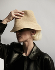 A person in a black leather jacket wears the JANESSA LEONE FELIX BUCKET raffia straw hat, tilted over their eyes and holding the brim with one hand against a plain, light background.