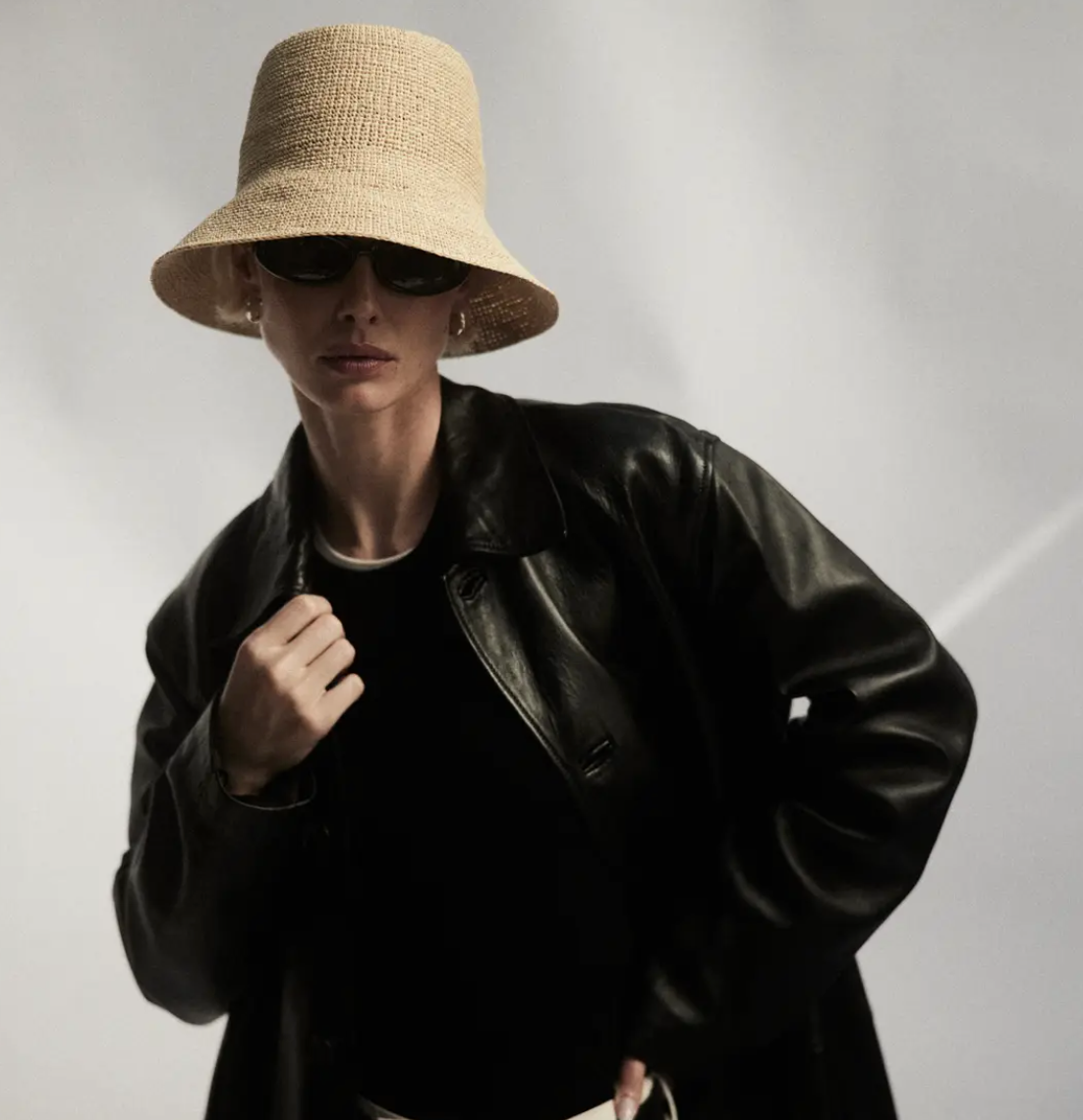 A stylish person wearing a Janessa Leone FELIX BUCKET NATURAL straw hat, sunglasses, and a black leather jacket, posing with attitude against an Arizona-themed background.
