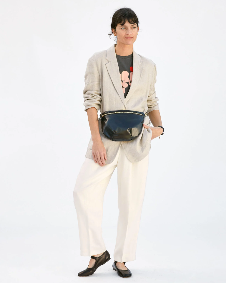 A woman in a stylish outfit featuring a beige blazer, white pants, and a Mickey Mouse t-shirt holds a Clare Vivier Grande Fanny Black shoulder bag, against an Arizona-inspired plain white background.