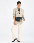 A woman in a stylish outfit featuring a beige blazer, white pants, and a Mickey Mouse t-shirt holds a Clare Vivier Grande Fanny Black shoulder bag, against an Arizona-inspired plain white background.