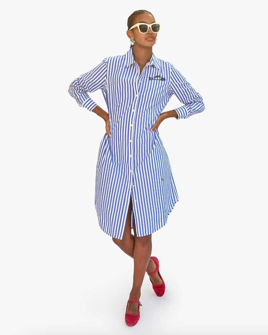 A woman wearing a Suzette Dress Blue & Cream Stripe w/ Sardine from Clare Vivier, with Arizona-style red shoes and sunglasses, posing confidently with hands on hips against a plain white background.