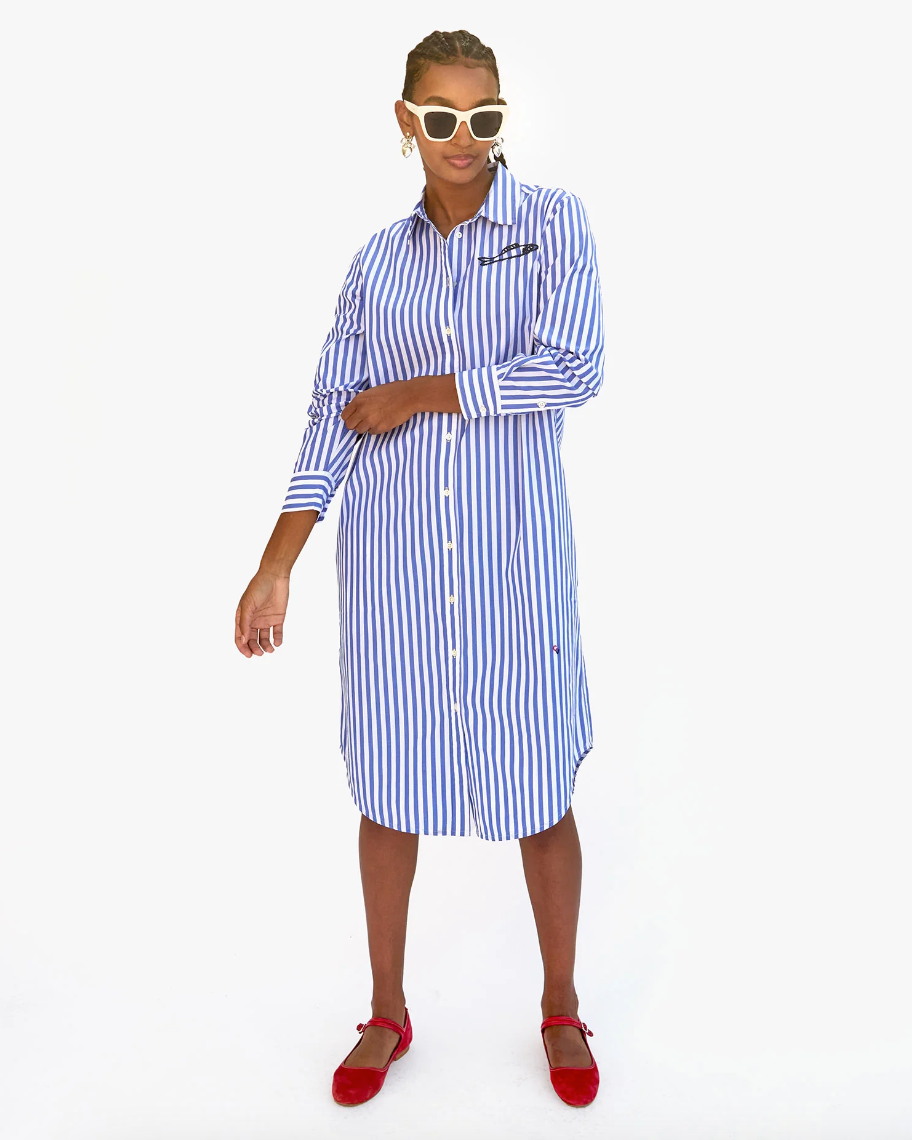 A woman wearing a Suzette Dress Blue &amp; Cream Stripe w/ Sardine by Clare Vivier, sunglasses, and red shoes stands confidently against a white background. She has short hair and her arms are crossed, exuding Arizona style.