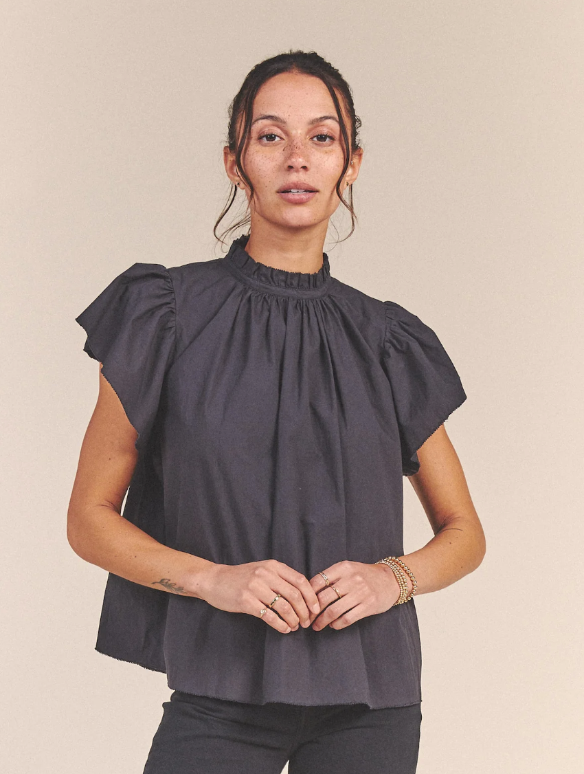 A woman in a CARLA HIGHNECK SHIRT BLACK POPLIN from Trovata stands against a plain background in her Scottsdale, Arizona bungalow, looking directly at the camera with her hands gently clasped in front of her.