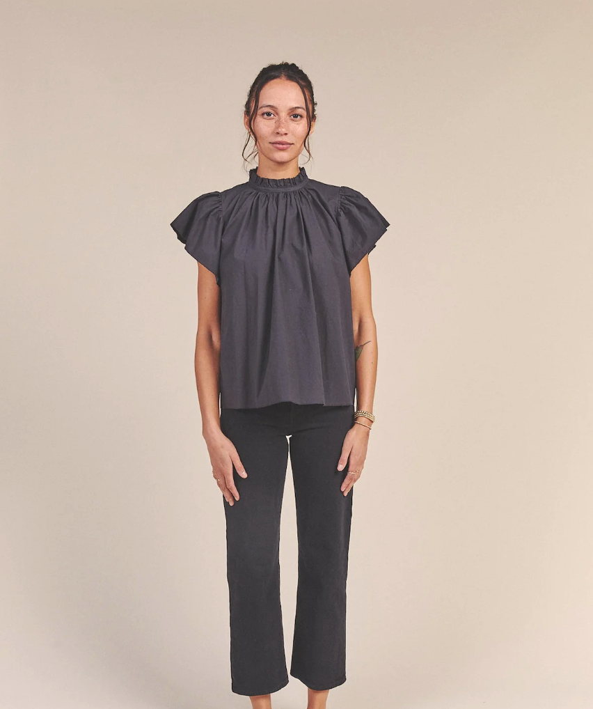 A woman standing against a neutral background, wearing a dark gray CARLA HIGHNECK SHIRT BLACK POPLIN with ruffled sleeves and black trousers, looking directly at the camera in Scottsdale, Arizona by Trovata.