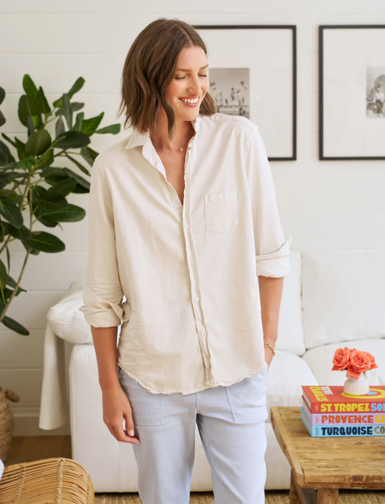 A woman smiling gently in a light-filled room, wearing a Frank & Eileen EILEEN Relaxed Button-Up Shirt in FAMOUS DENIM Vintage White and light blue pants, standing near a soft beige couch in an Arizona-style bungalow with books and a small orange flower on the coffee table.