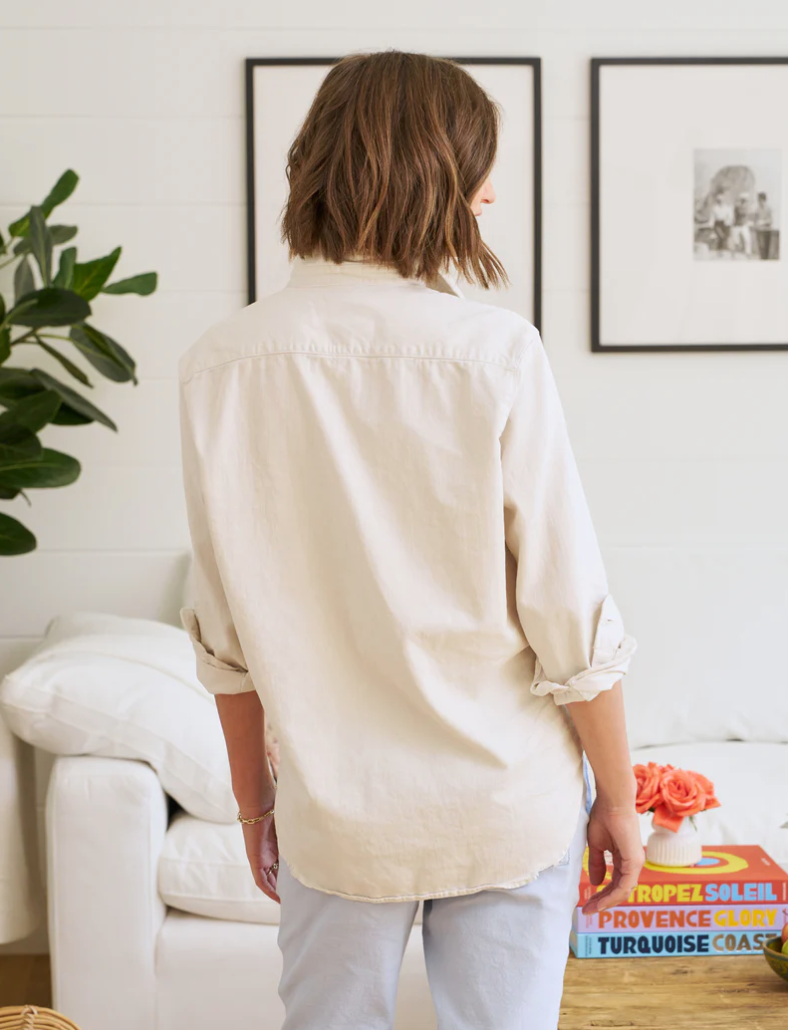 A woman stands in a bright living room of a bungalow, viewed from behind. She wears a Frank & Eileen Relaxed Button-Up Shirt in Vintage White and FAMOUS DENIM faded blue jeans, her brown hair cut in a bob. A white couch and potted plant are visible nearby.