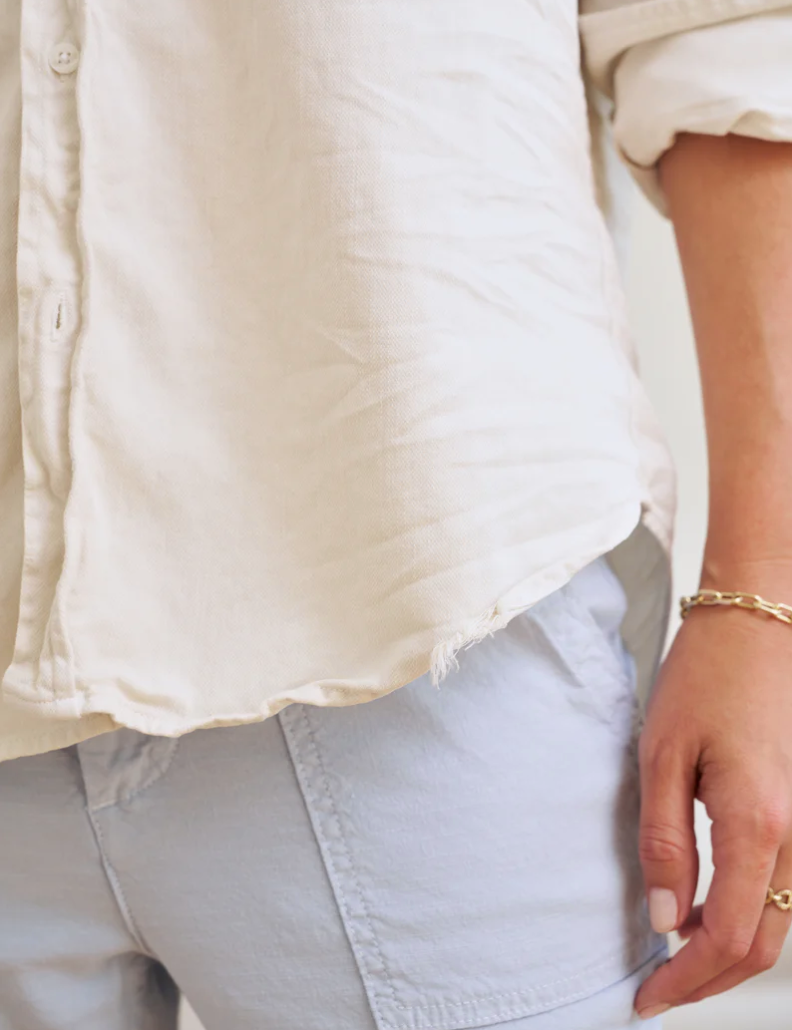 Close-up of a person wearing a Frank & Eileen EILEEN Relaxed Button-Up Shirt in FAMOUS DENIM Vintage White and blue denim shorts, with a hand casually placed near the pocket area. The focus is on the clothing style and relaxed fit.
