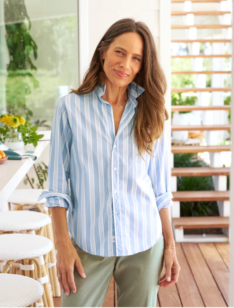 A woman with long brown hair smiles softly, wearing a Frank & Eileen BARRY Tailored Button-Up Shirt SUPERLUXE Slate Blue Stripe and green pants, standing in a brightly lit bungalow with plants in the background.