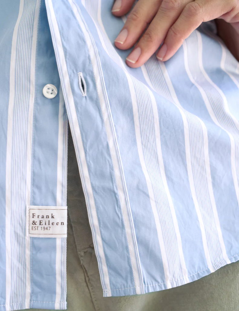 A close-up of a Frank & Eileen BARRY Tailored Button-Up Shirt SUPERLUXE Slate Blue Stripe with the brand tag "Frank & Eileen EST 1947" visible inside, styled elegantly in an Arizona bungalow, and a hand gently pulling the shirt open.