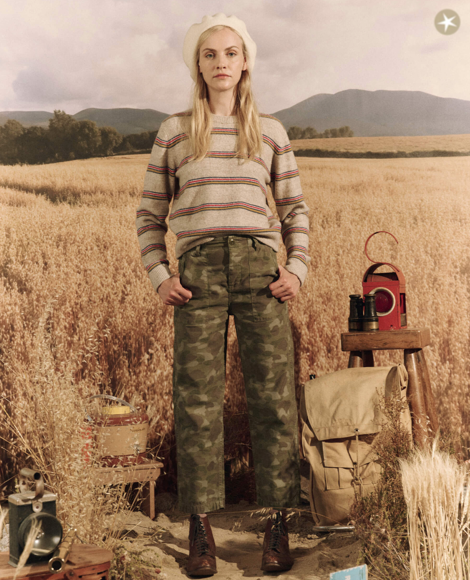 A woman in a striped sweater, The Admiral Pant from The Great Inc., and combat boots stands in a wheat field with backpacking gear and a red lantern, looking pensively at the camera. Her outfit reflects an Arizona style influence.