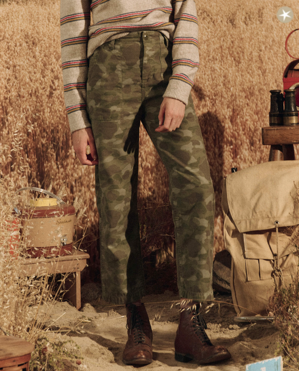 A person wearing a striped sweater and The Admiral Pant in DESERT CAMO stands in a rustic outdoor setting with tall grass, with hiking boots and a backpack nearby.