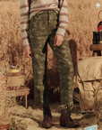 A person wearing a striped sweater and The Admiral Pant in DESERT CAMO stands in a rustic outdoor setting with tall grass, with hiking boots and a backpack nearby.