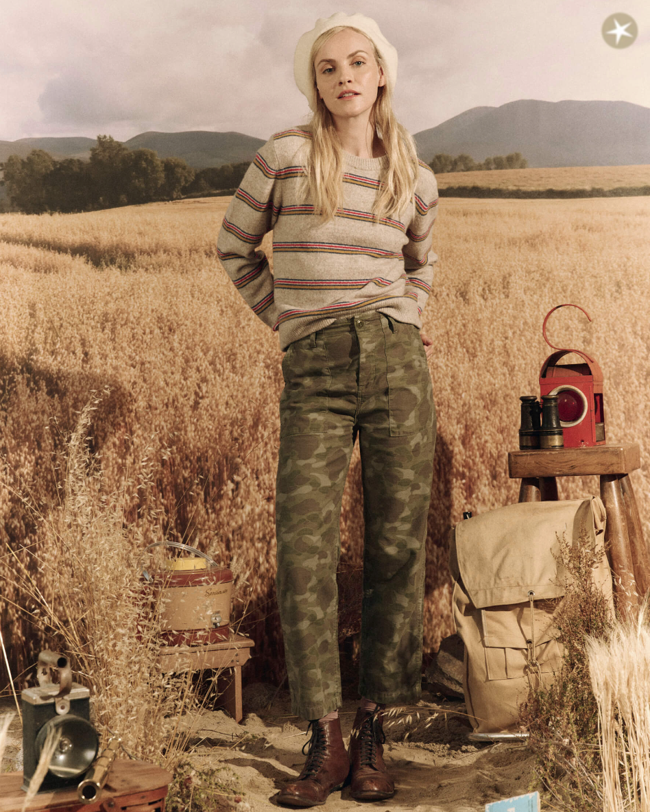 A woman in a striped sweater and The Admiral Pant. DESERT CAMO stands confidently in an Arizona wheat field, with a vintage lantern, backpack, and small items on a wooden stool beside her from The Great Inc.