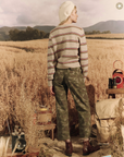 A person stands in a golden wheat field in Arizona, looking into the distance. They wear a striped sweater, The Admiral Pant in DESERT CAMO by The Great Inc., and brown boots, with a white hat. Various rustic camping gear is scattered nearby.