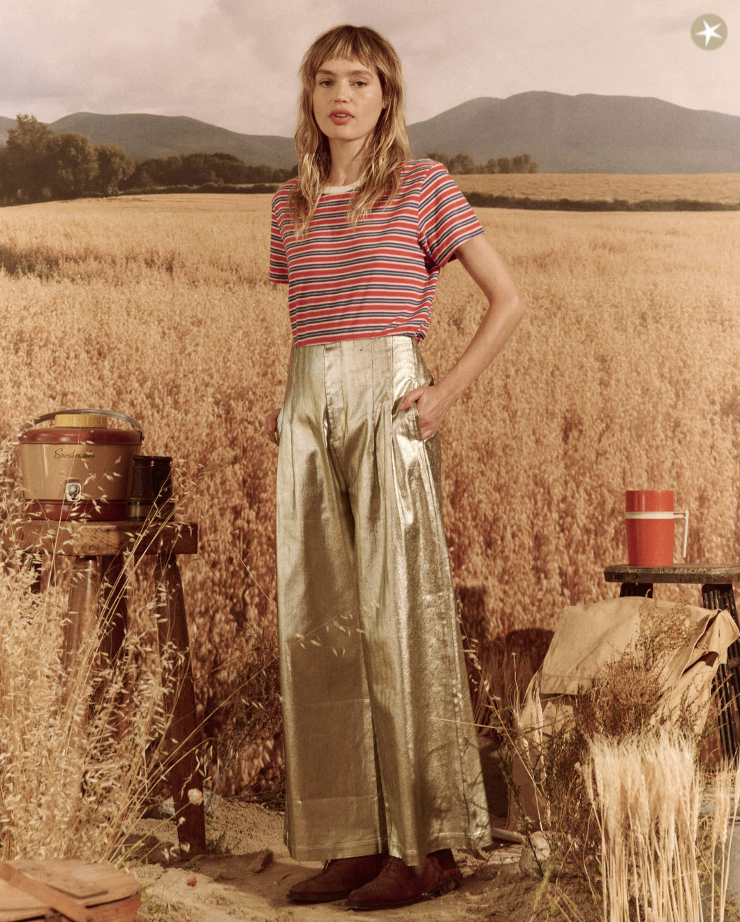 A woman stands in a wheat field wearing The Great Inc.'s Sculpted Trousers, a red and blue striped T-shirt, and brown shoes. A rustic barrel and can are visible in the background under a cloudy sky, adding to the Arizona-style ambiance.