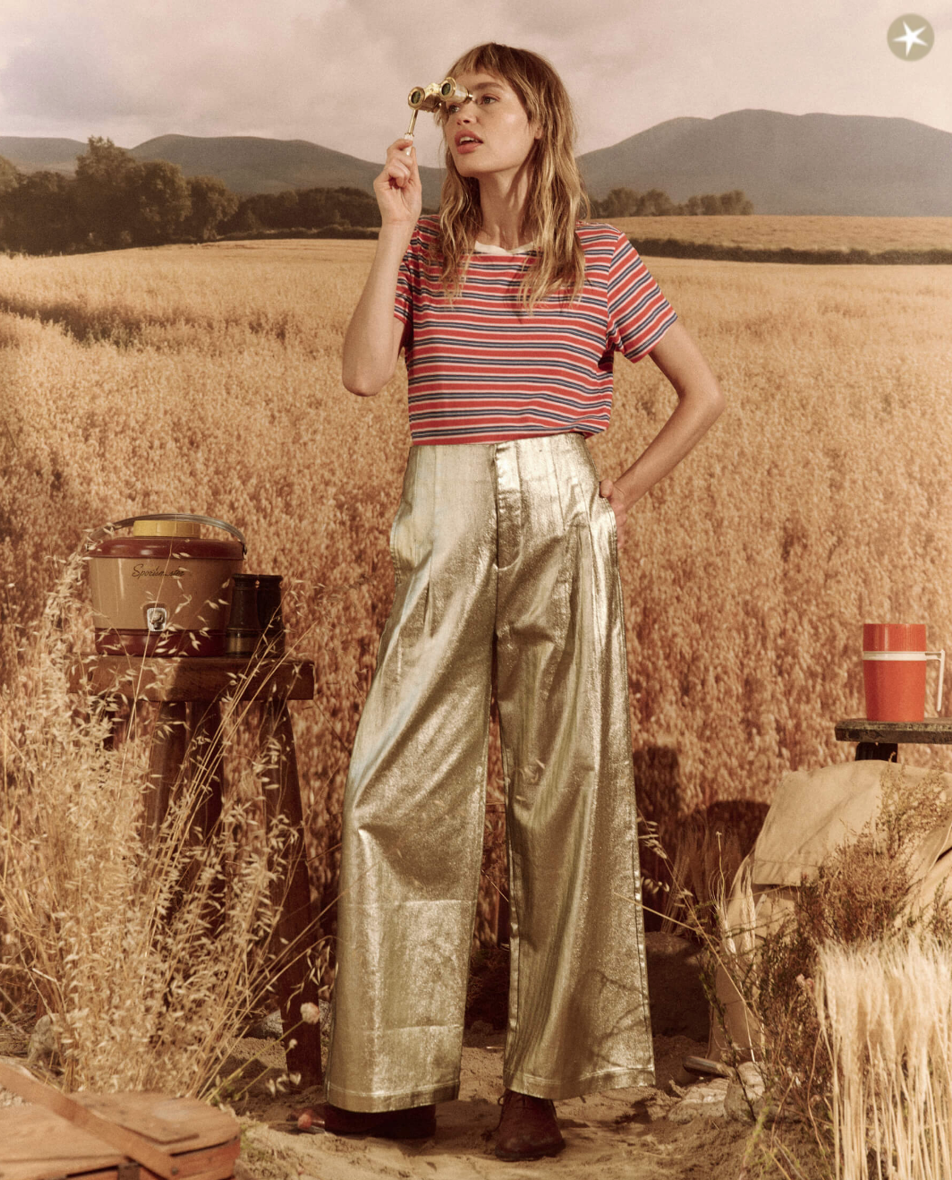 A woman with a short haircut, wearing a striped red and blue top and The Great Inc.'s Sculpted Trouser, stands in an Arizona wheat field, looking through a monocle with a thoughtful expression.