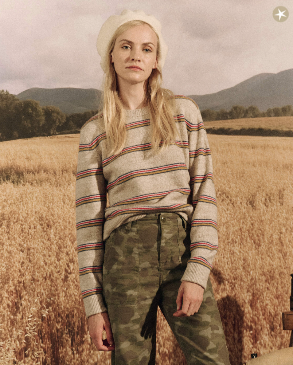 A young woman in a striped sweater and camo pants stands in a wheat field, wearing a white beret with The Shrunken Pullover. WATERFRONT STRIPE style, with mountains in the background by The Great Inc.