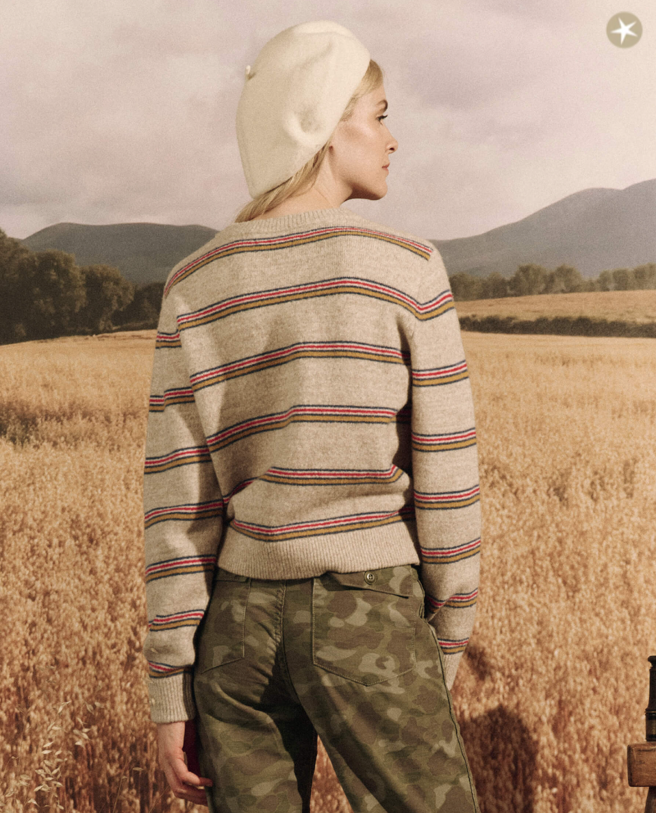 A woman in a Waterfront Stripe Shrunken Pullover from The Great Inc. stands in a golden wheat field, looking over her shoulder, with distant hills under a soft Arizona sky.