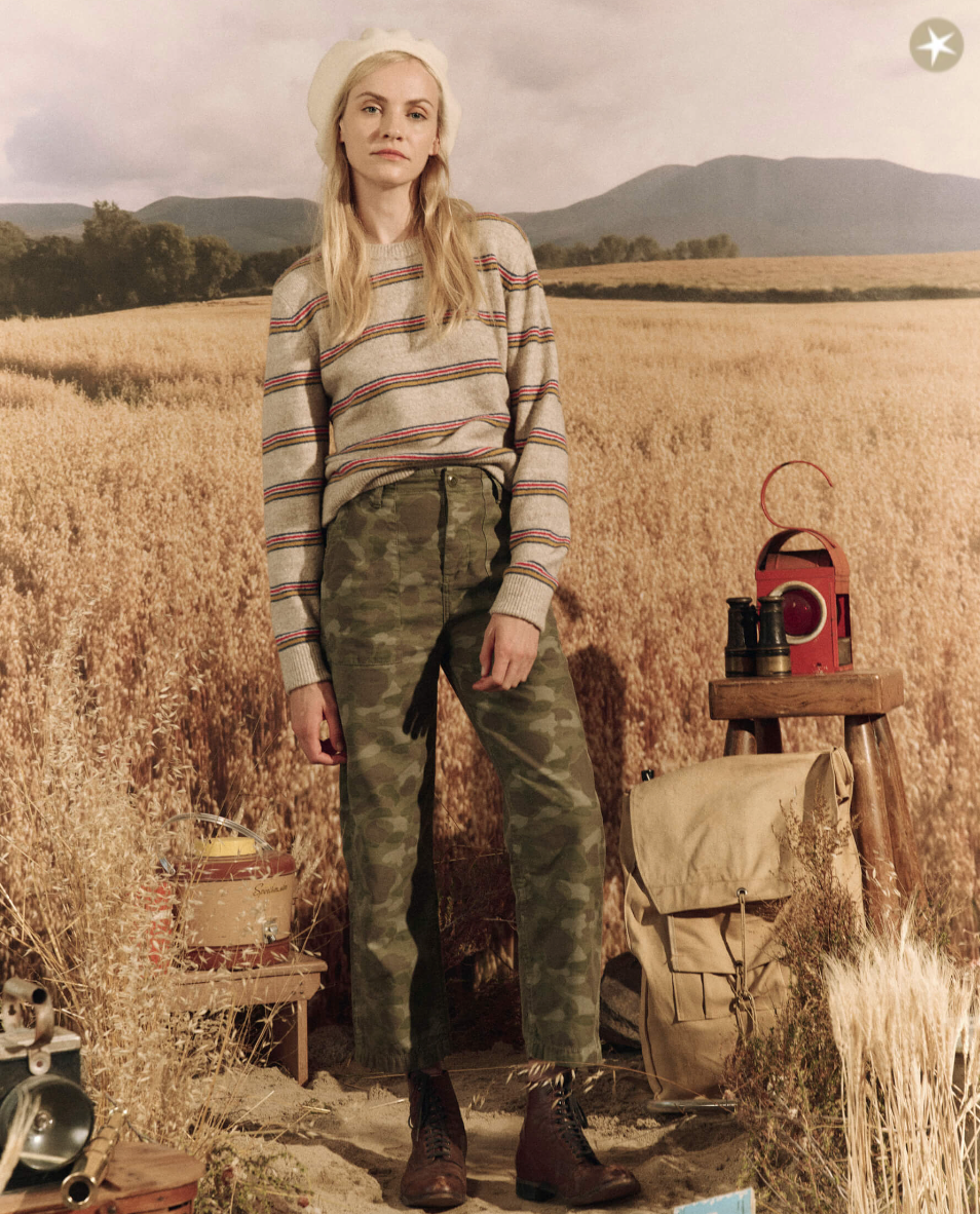 A woman stands in a golden wheat field, wearing The Shrunken Pullover from WATERFRONT STRIPE brand, camo pants, and brown boots. Beside her is a table with a red lantern and canvas bag. Hills and an Arizona-style cloudy sky are in the background.
