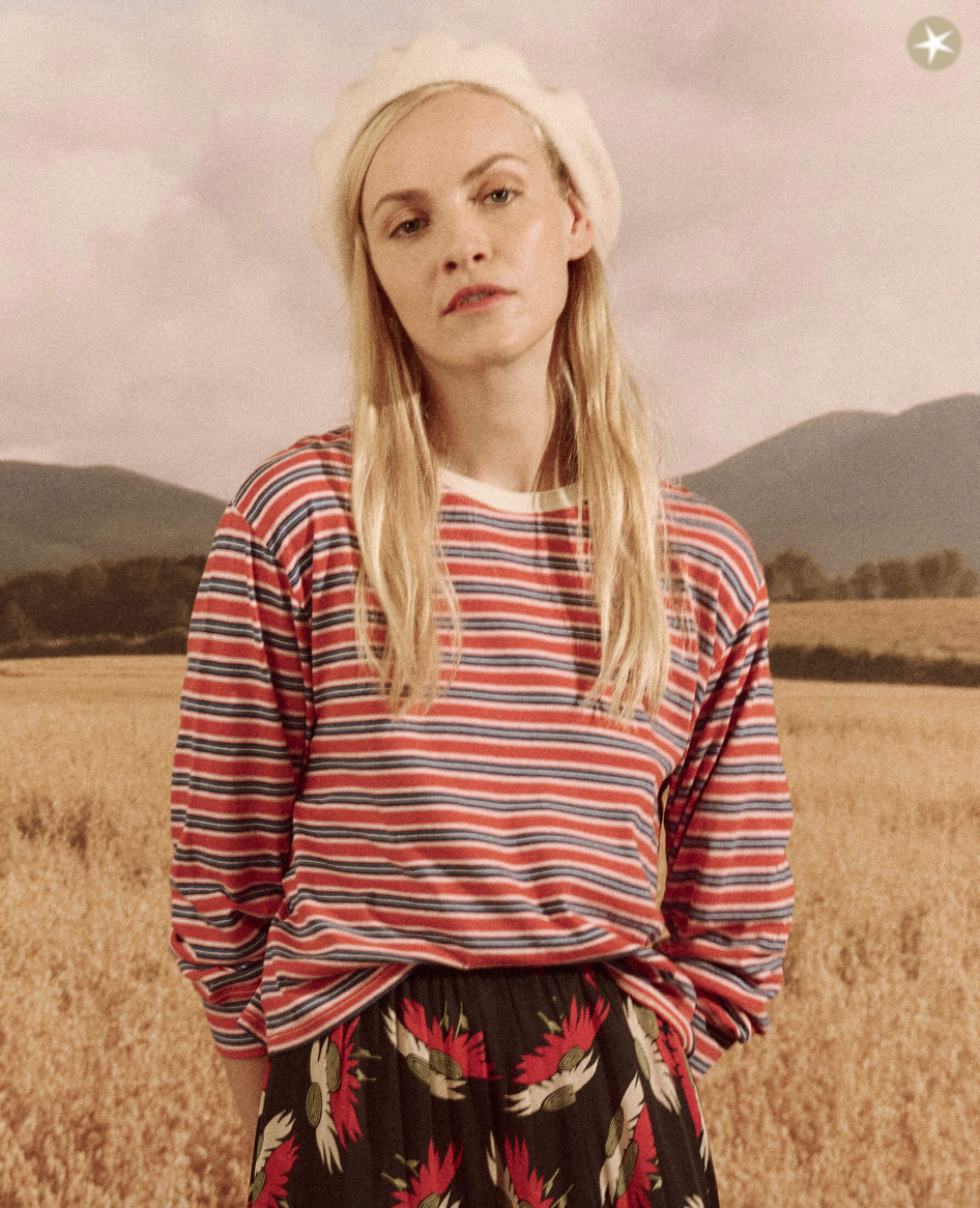 A woman wearing The Campus Crew CAMPERVAN STRIPE shirt and a floral skirt stands in a wheat field, with mountains in the background and a clear Arizona sky. She has a thoughtful expression and wears a white beret.