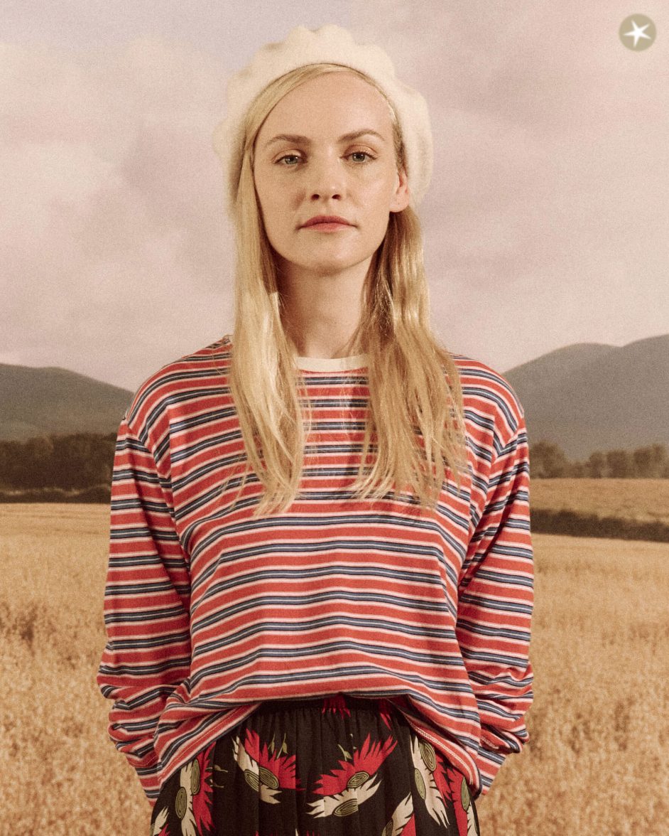A woman with blond hair sporting a white beret and a striped long-sleeve shirt stands in front of a pastoral field with distant mountains under a subdued Arizona sky wearing The Great Inc.'s The Campus Crew CAMPERVAN STRIPE.