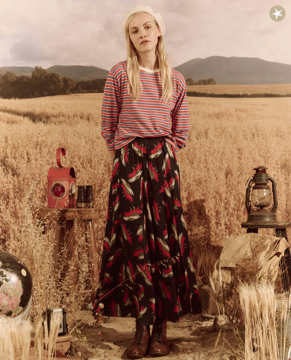 A woman in The Great Inc.'s CAMPERVAN STRIPE top and floral skirt stands in a golden wheat field with vintage items like a camera and lantern around her. Distant hills under a cloudy sky form the background, evoking the Arizona style.