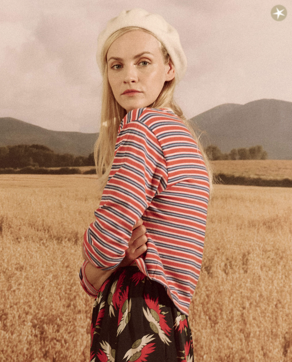 A woman in a CAMPERVAN STRIPE top and floral skirt with a white beret looks over her shoulder in a golden wheat field, with distant mountains under an Arizona hazy sky by The Great Inc.