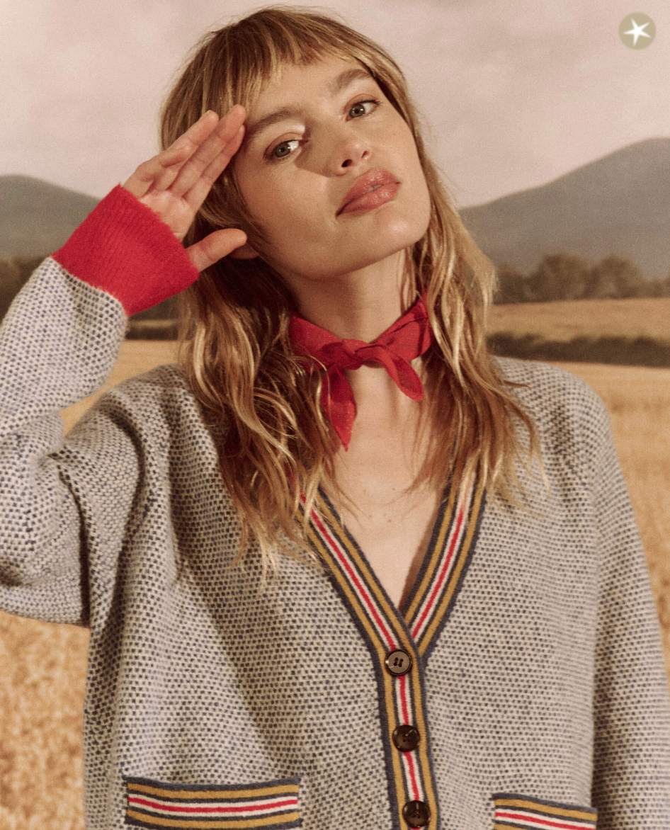 A woman with a playful expression shields her eyes with her hand, standing in a wheat field. She wears The Varsity Cardigan by COASTLINE, a red scarf tied at her neck, and has light brown hair with bangs.