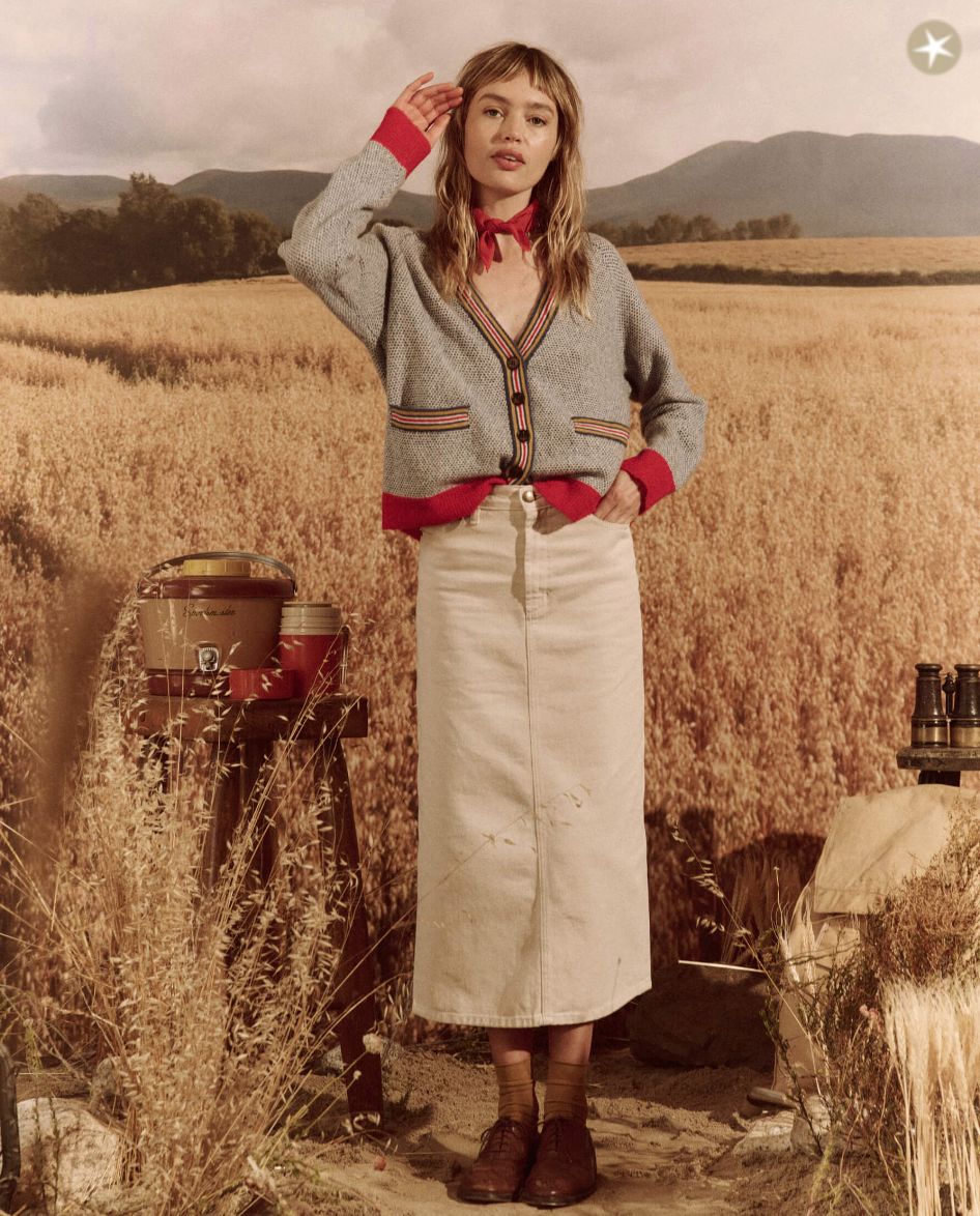 A woman stands in a field of tall grass, wearing The Varsity Cardigan by COASTLINE over a striped shirt, a beige skirt, red wristbands, and brown shoes. A rustic drum and plants surround her, evoking the style of an Arizona landscape.