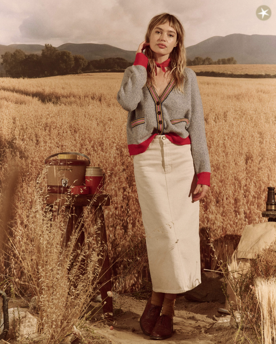 A woman stands in a field at sunset, wearing a colorful Arizona-style blouse, The Varsity Cardigan from COASTLINE, and long white skirt, touching her cheek thoughtfully. Countryside backdrop with mountains and agricultural machinery.