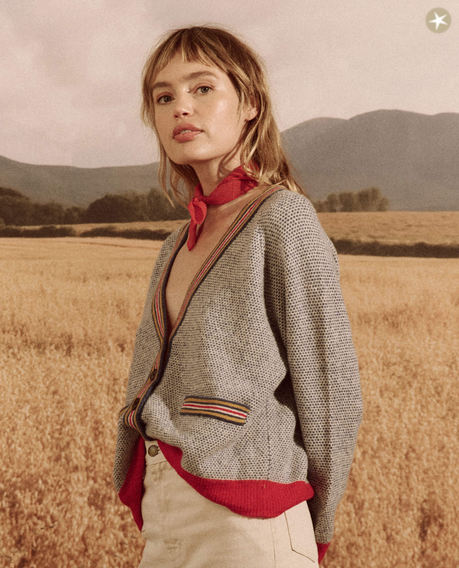 A woman stands in a field, wearing The Varsity Cardigan by COASTLINE over a red blouse with a necktie, gazing gently at the camera. Golden fields and distant Arizona mountains form the backdrop.