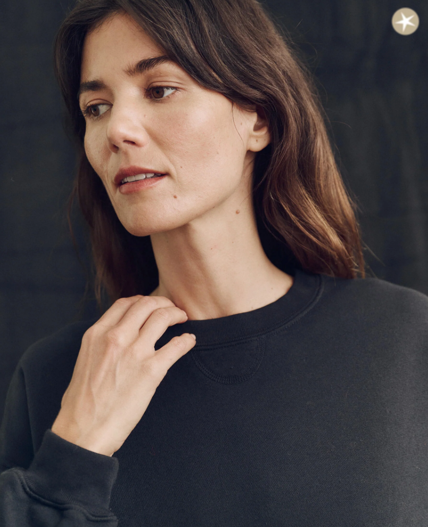 A close-up portrait of a woman with shoulder-length brown hair, looking to the side thoughtfully, wearing The League Sweatshirt ALMOST BLACK against a black backdrop in Scottsdale, Arizona by The Great Inc.
