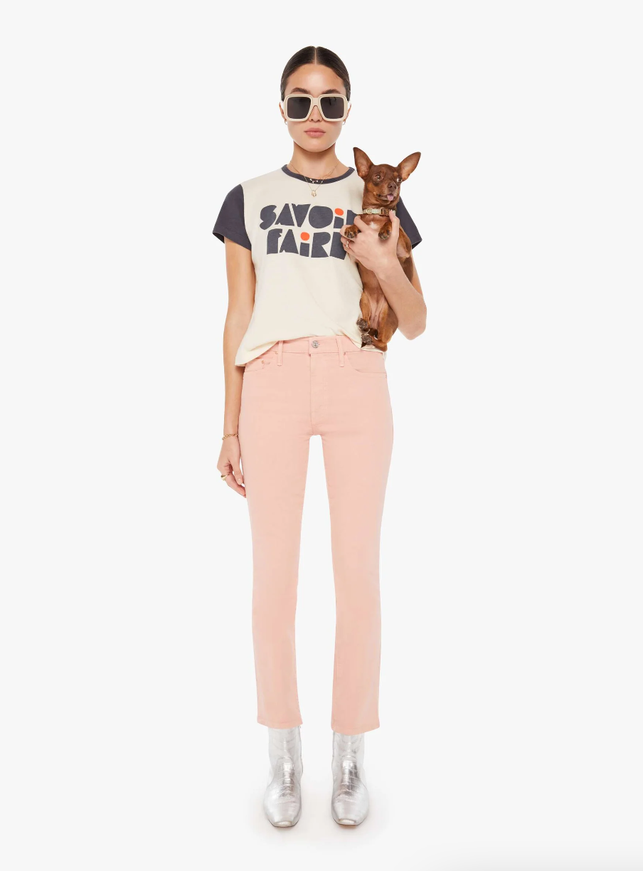 A woman in stylish attire, wearing a Mother's Goodie Goodie ringer Savoir Faire tee, pink pants, and silver boots, holds a small brown dog. She accessorizes with large sunglasses and poses against a bungalow in Scottsdale, Arizona.