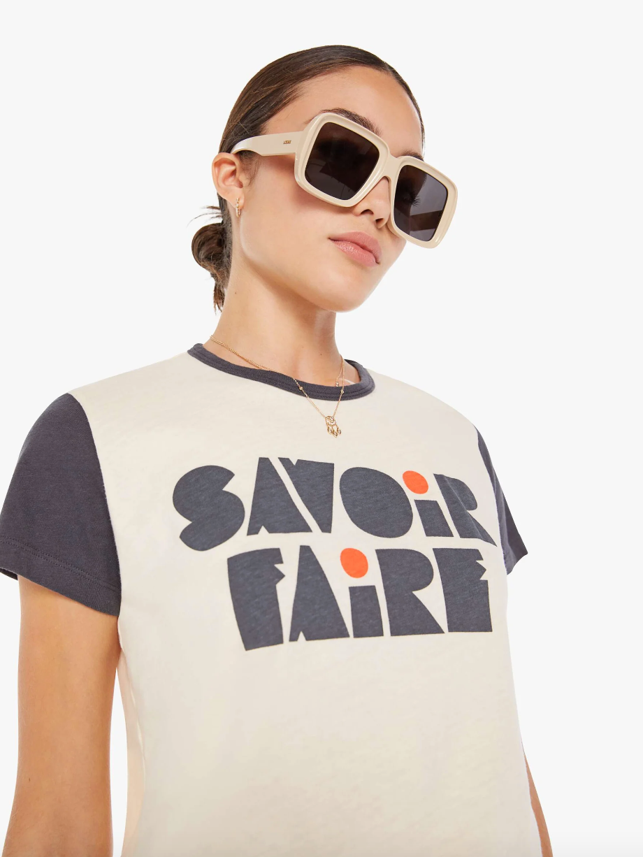 A woman wearing large white sunglasses and a t-shirt with "the Goodie Goodie ringer Savoir Faire" printed on it in stylized letters, posing against a plain background in Scottsdale, Arizona by Mother.