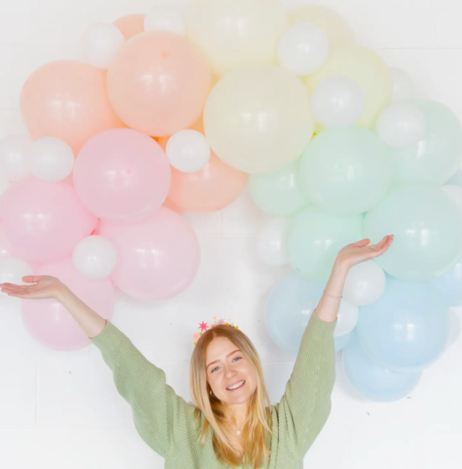 A woman with a joyful expression throws her arms open among a cluster of Faire Balloon Arch Kit Party Dec balloons in shades of pink, peach, yellow, and blue in an Arizona bungalow.