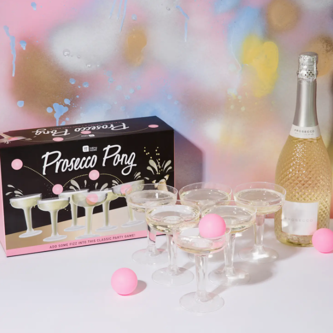 A party setup at a Scottsdale, Arizona bungalow featuring a box labeled &quot;Prosecco Pong Drinking Game&quot; by Faire, several champagne glasses arranged for a game, pink balls, and a bottle of prosecco against a colorful backdrop.
