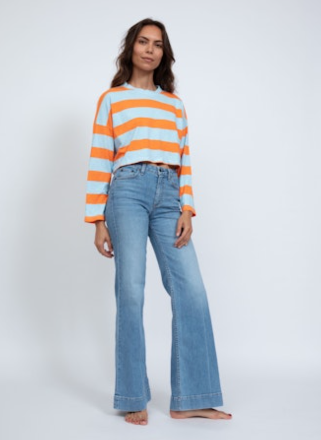 A person stands confidently in an ASKK printed cropped long sleeve and light blue flare jeans, barefoot against a plain white background, capturing the relaxed vibe of Los Angeles.