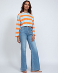 A person stands confidently in an ASKK printed cropped long sleeve and light blue flare jeans, barefoot against a plain white background, capturing the relaxed vibe of Los Angeles.